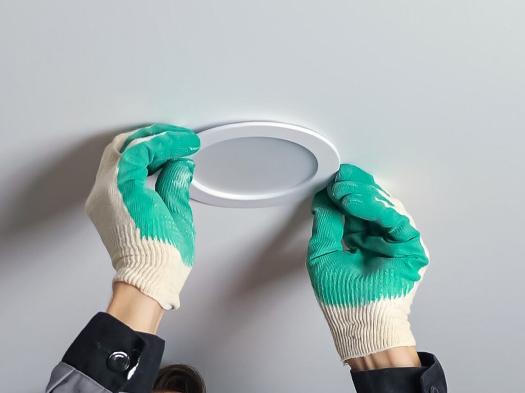 Professional short-haired electrician in work gloves and grey jacket installs light on ceiling standing on ladder during apartment renovation.