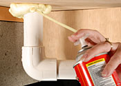 Someone using spray foam to seal pipes under a sink